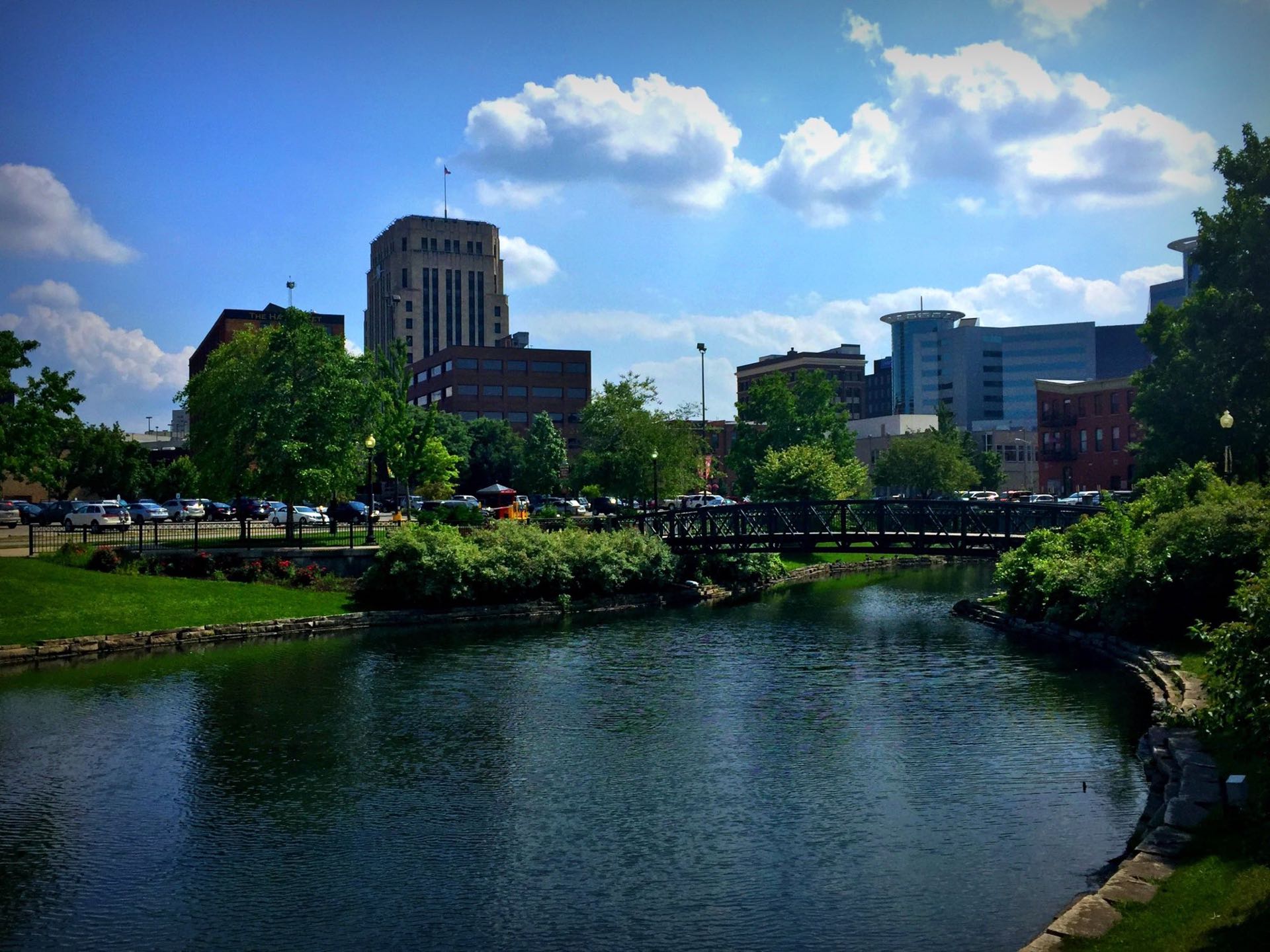 The downtown Kalamazoo skyline seen across the creek, with a footbridge and a park path along the water