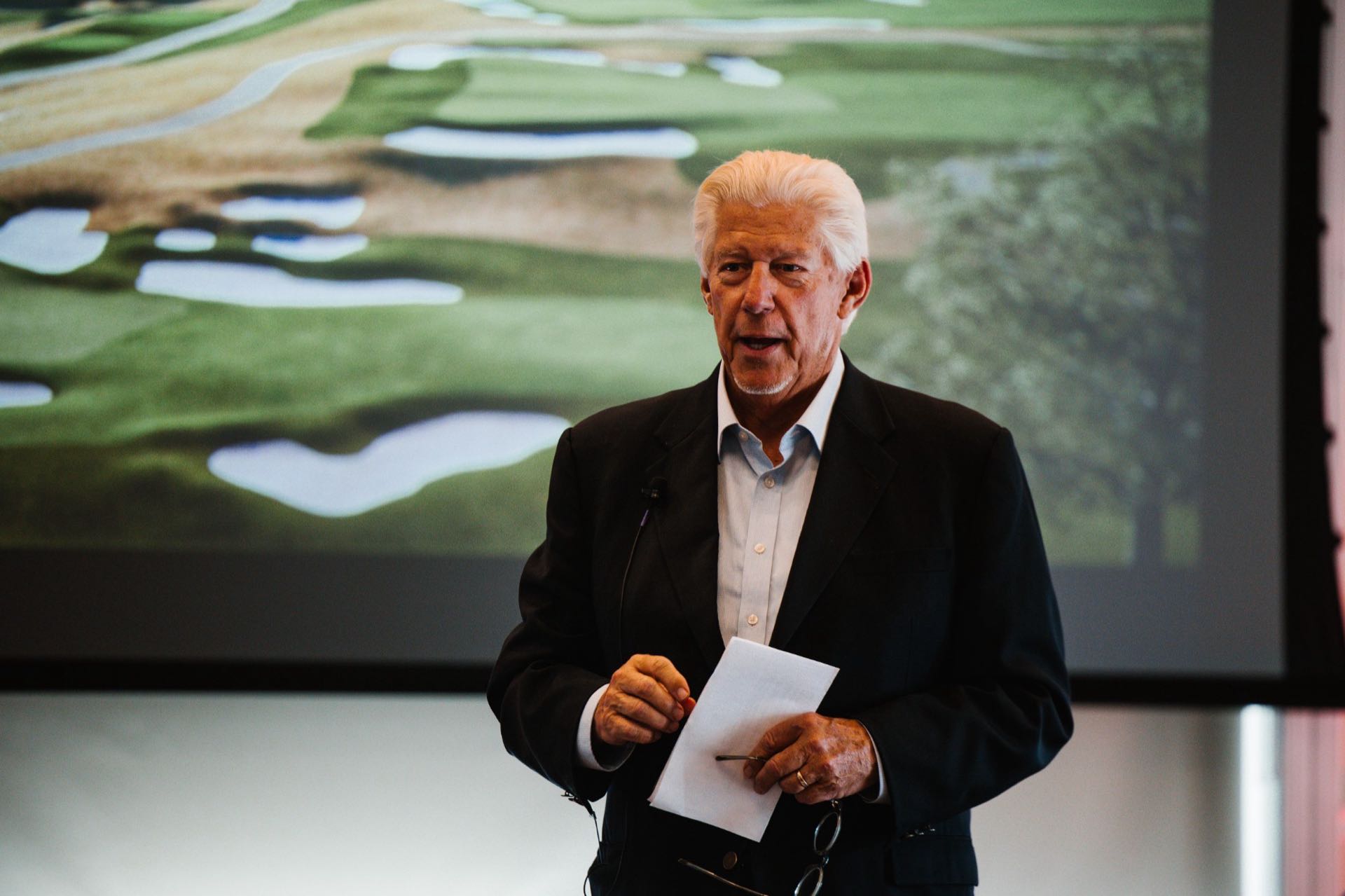 William D. Johnston speaking to a seated audience, a rendering of the new championship golf course on the screen behind him